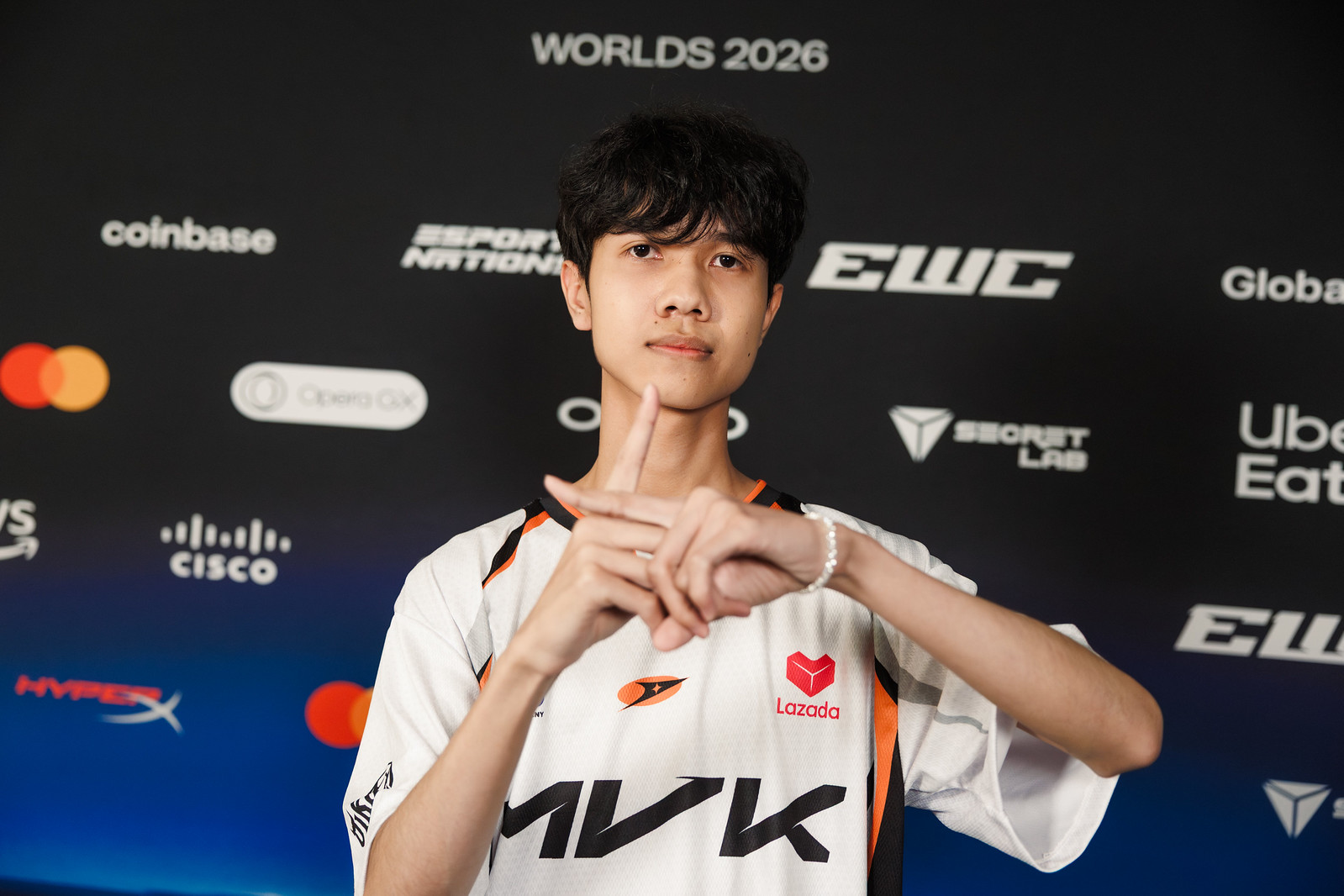 Một tuyển thủ MVK Esports trong áo đấu trắng cam bắt chéo hai tay thành chữ X trước phông nền Worlds 2026